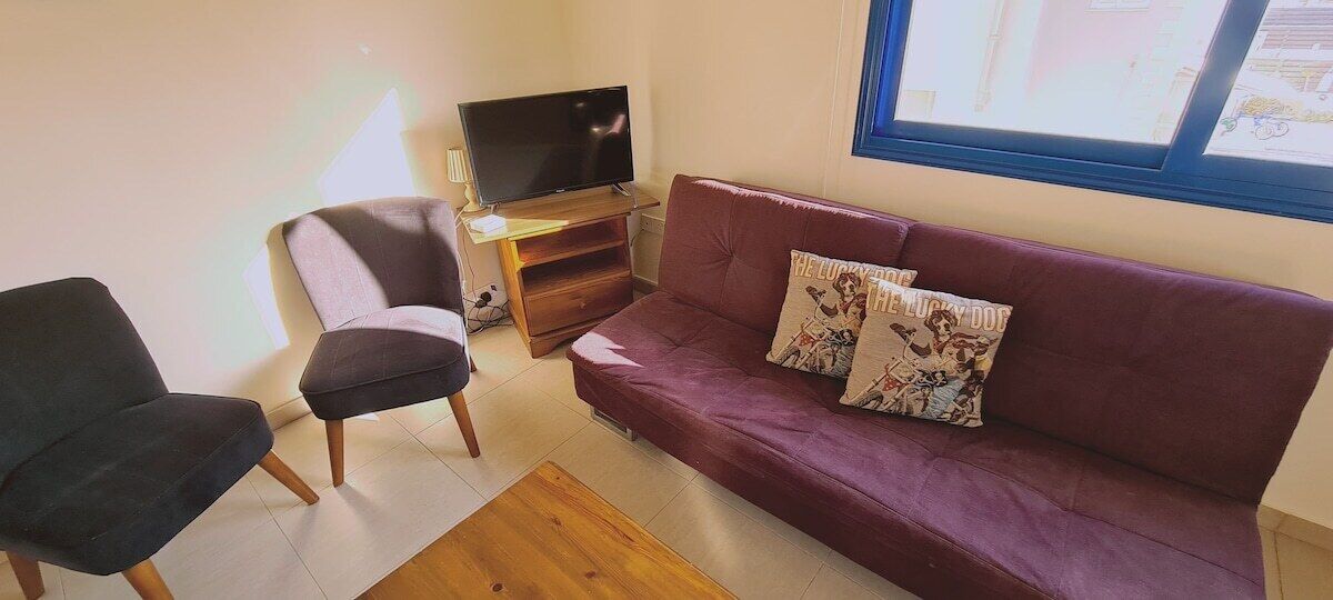 Un salon comprenant un canapé, deux fauteuils, un téléviseur posé sur un meuble en bois et une fenêtre avec des stores.