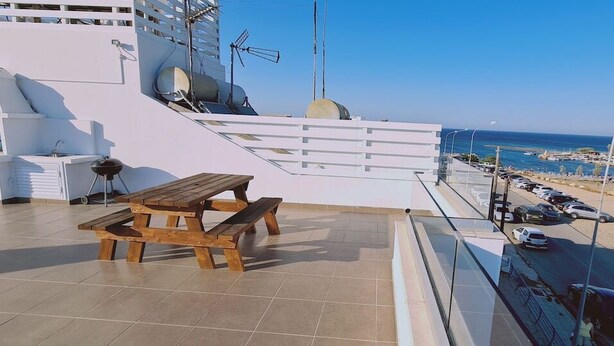 Une terrasse sur le toit, avec une table et des chaises en bois, un barbecue et une vue sur la mer et les voitures garées.