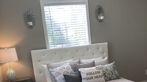 A bedroom with a tufted headboard, a bedside table with a lamp, a window with blinds, and decorative wall mirrors. - Comfy and Cozy Centrally located in Houston!! (Houston)