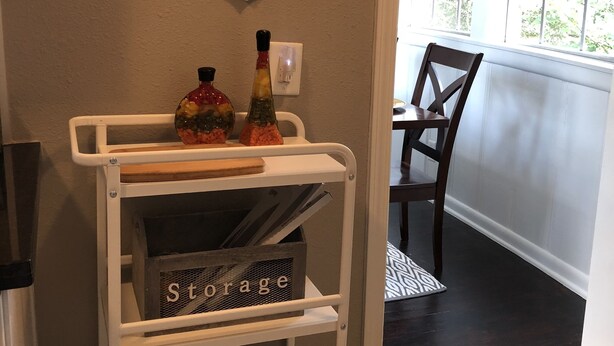 A white storage cart with shelves, a 'Storage' sign, and decorative bottles. - Comfy and Cozy Centrally located in Houston!! (Houston)