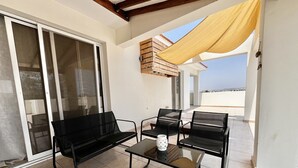 Un balcon avec un canapé noir, deux chaises noires, une table en verre et un auvent jaune.