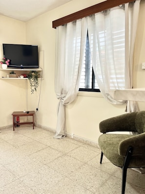 Ein Zimmer mit Fernseher auf einem Regal, einem Fenster mit Vorhängen, einem kleinen Tisch und einem Stuhl.