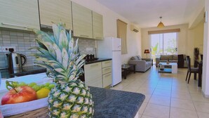 Eine moderne Küche mit Kühlschrank, Herd und Spüle. Ein Essbereich mit Tisch und Stühlen ist vorhanden. Auf der Arbeitsfläche sind eine große Ananas und eine Obstschale zu sehen.
