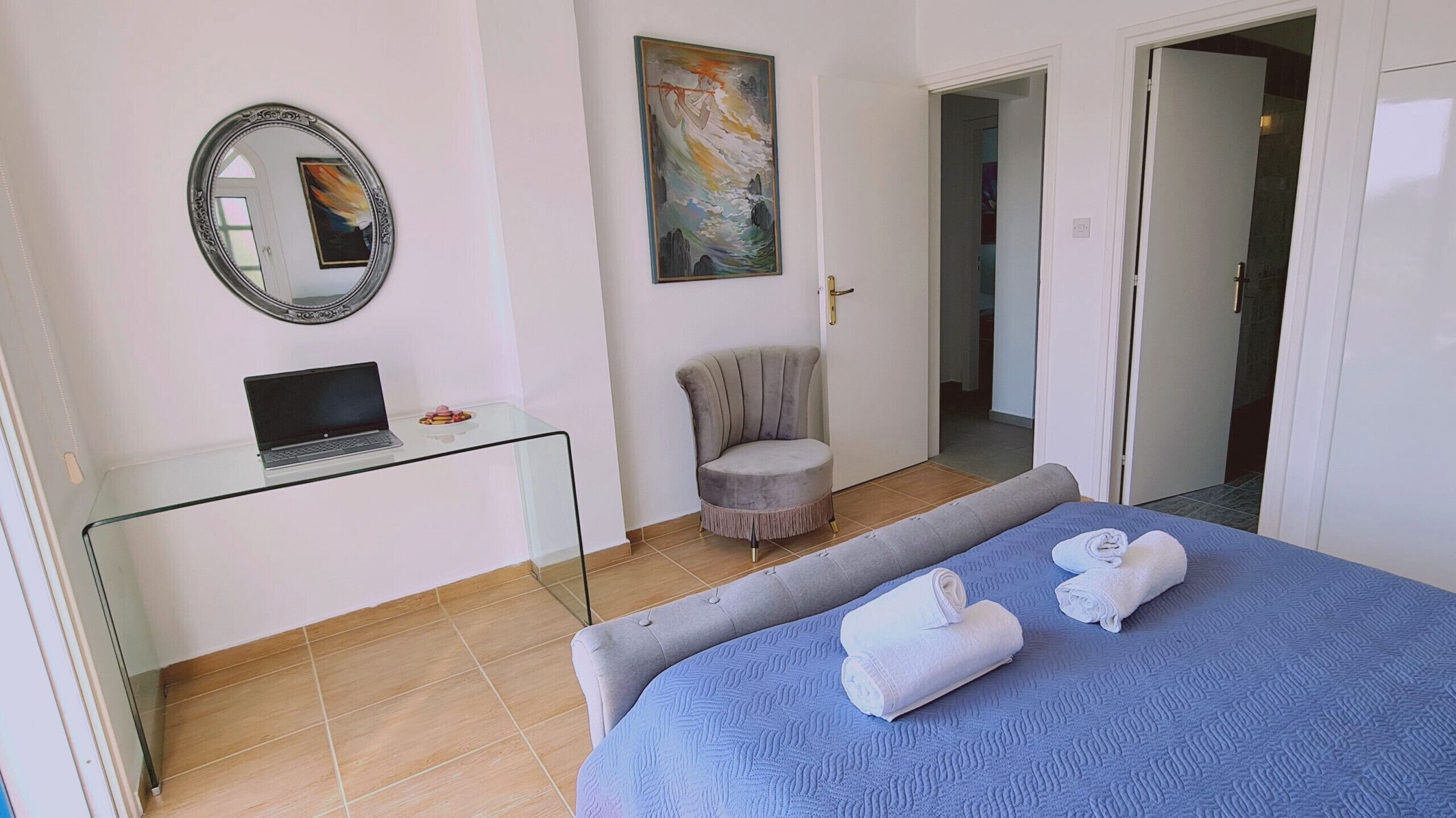 A bedroom with a bed, a desk with a laptop, a chair, and a door leading to another room.
