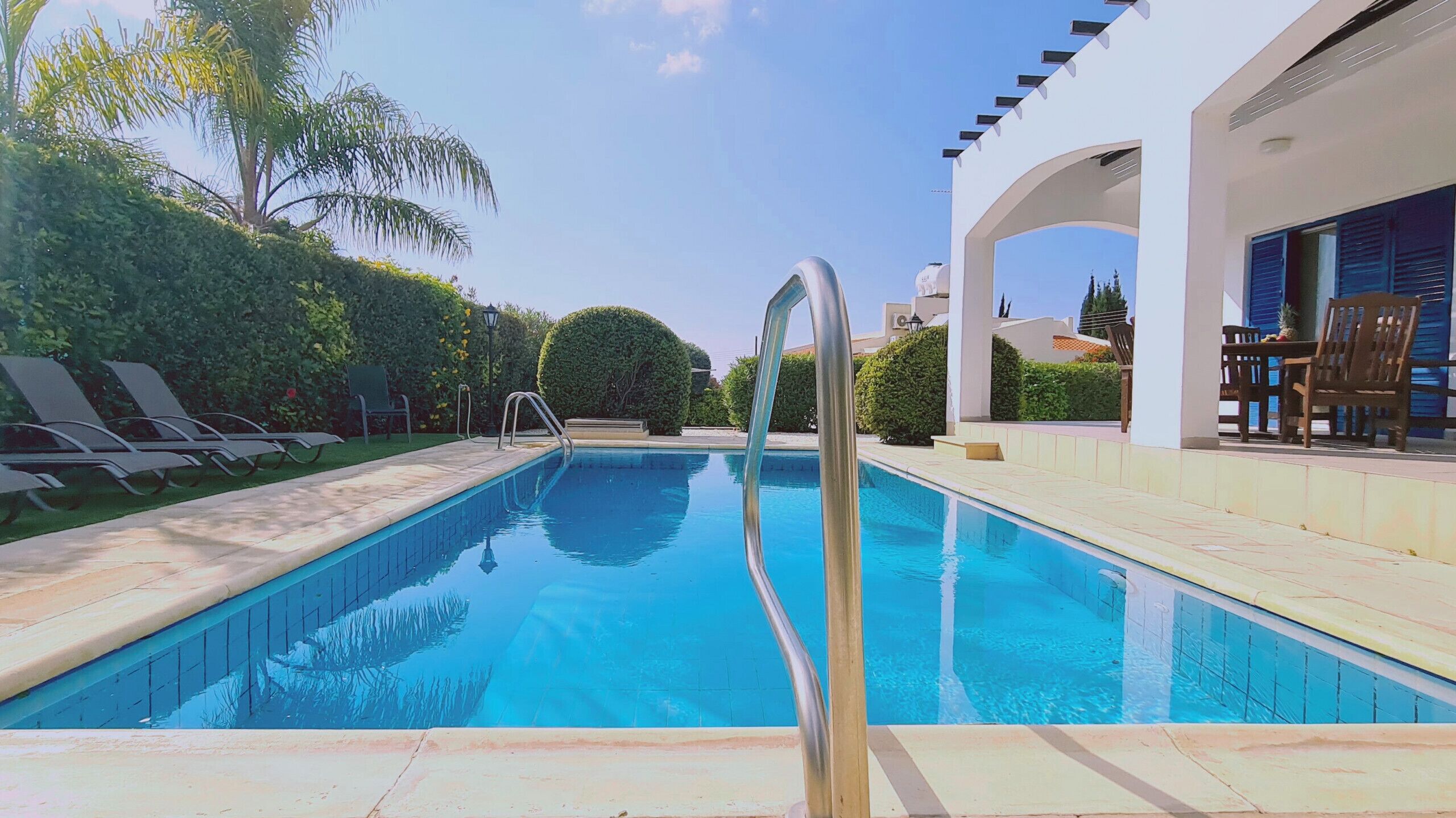 Una piscina con una escalera metálica, rodeada de sillas de descanso y un edificio blanco con un patio cubierto.
