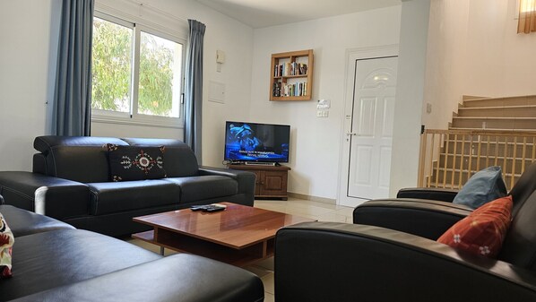 A living room with a black leather sofa, a wooden coffee table, and a television on a wooden stand. There is a bookshelf with books and a door in the background.