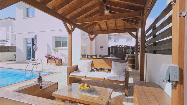 A covered outdoor seating area with a pool, wooden furniture, and a table with fruit. - STAY Villa Anemone (Protaras)