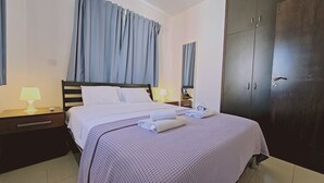 A neatly made bed with a white duvet and a purple knitted blanket, two bedside tables with lamps, and a dark wooden wardrobe. - STAY Villa Anemone (Protaras)