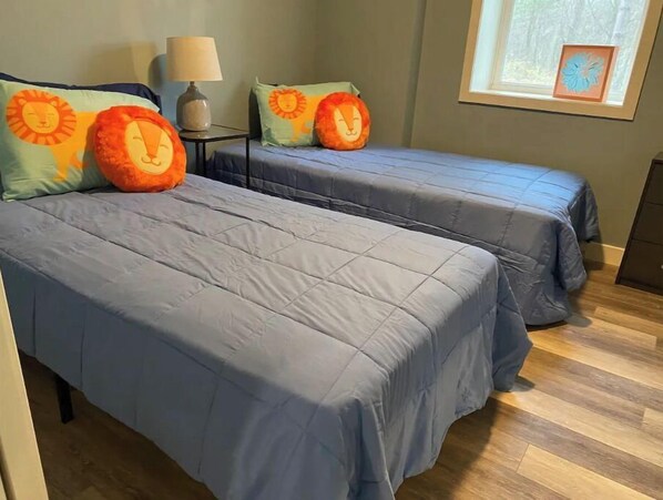 A room with two beds, each with a blue bedspread and orange lion-themed pillows. There is a small table with a lamp between the beds.