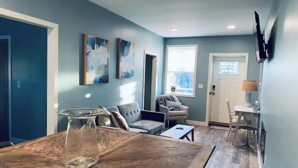 Una sala de estar con una mesa de comedor de madera, un sofá gris y una silla. Hay dos cuadros en la pared, una ventana y una puerta.