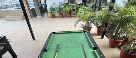 Una mesa de billar con bolas dispuestas en una pirámide, rodeada de plantas en macetas, en un espacio interior con vista a un paisaje urbano.