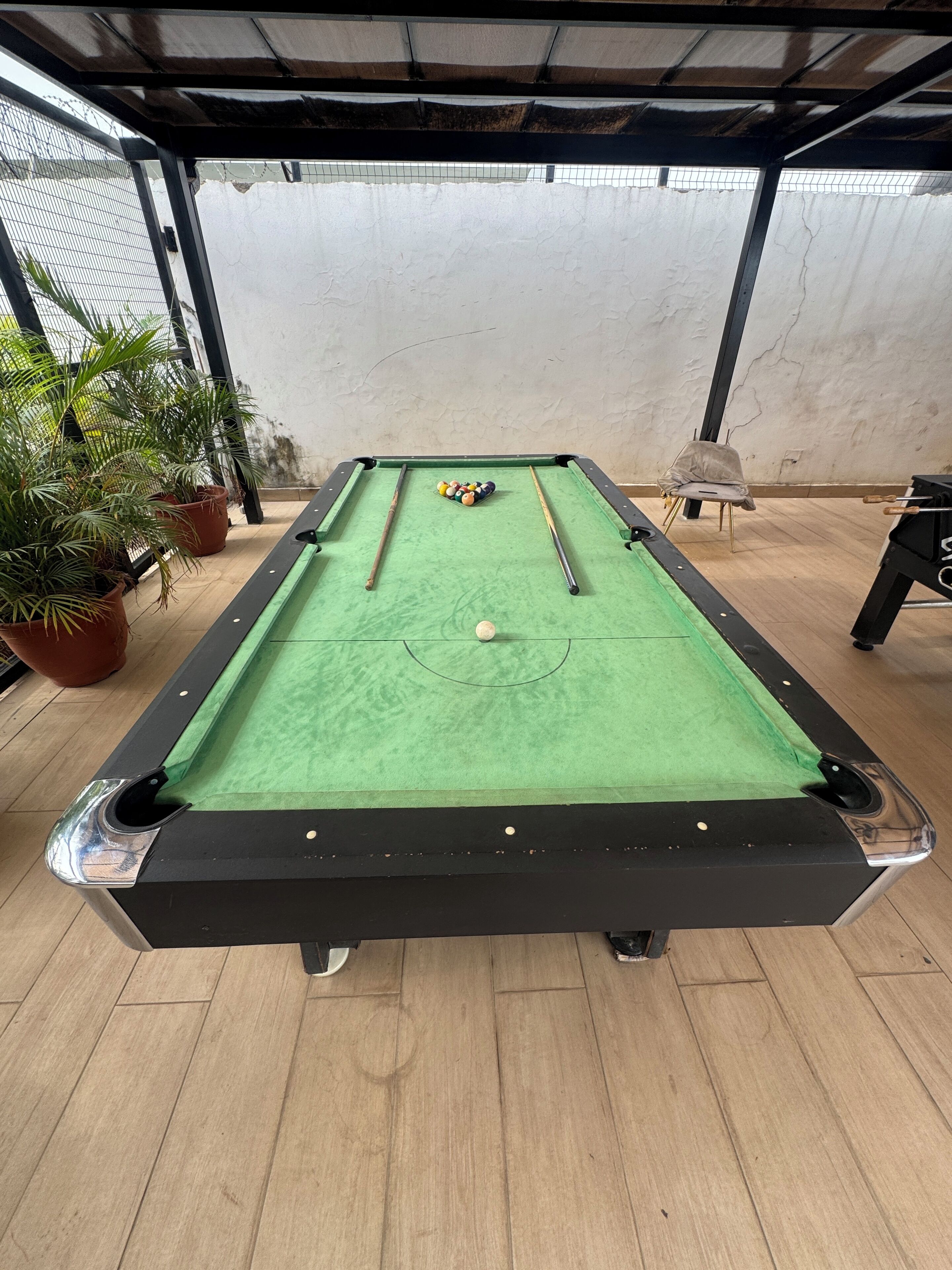 A pool table with a green felt surface, surrounded by potted plants and a chair.