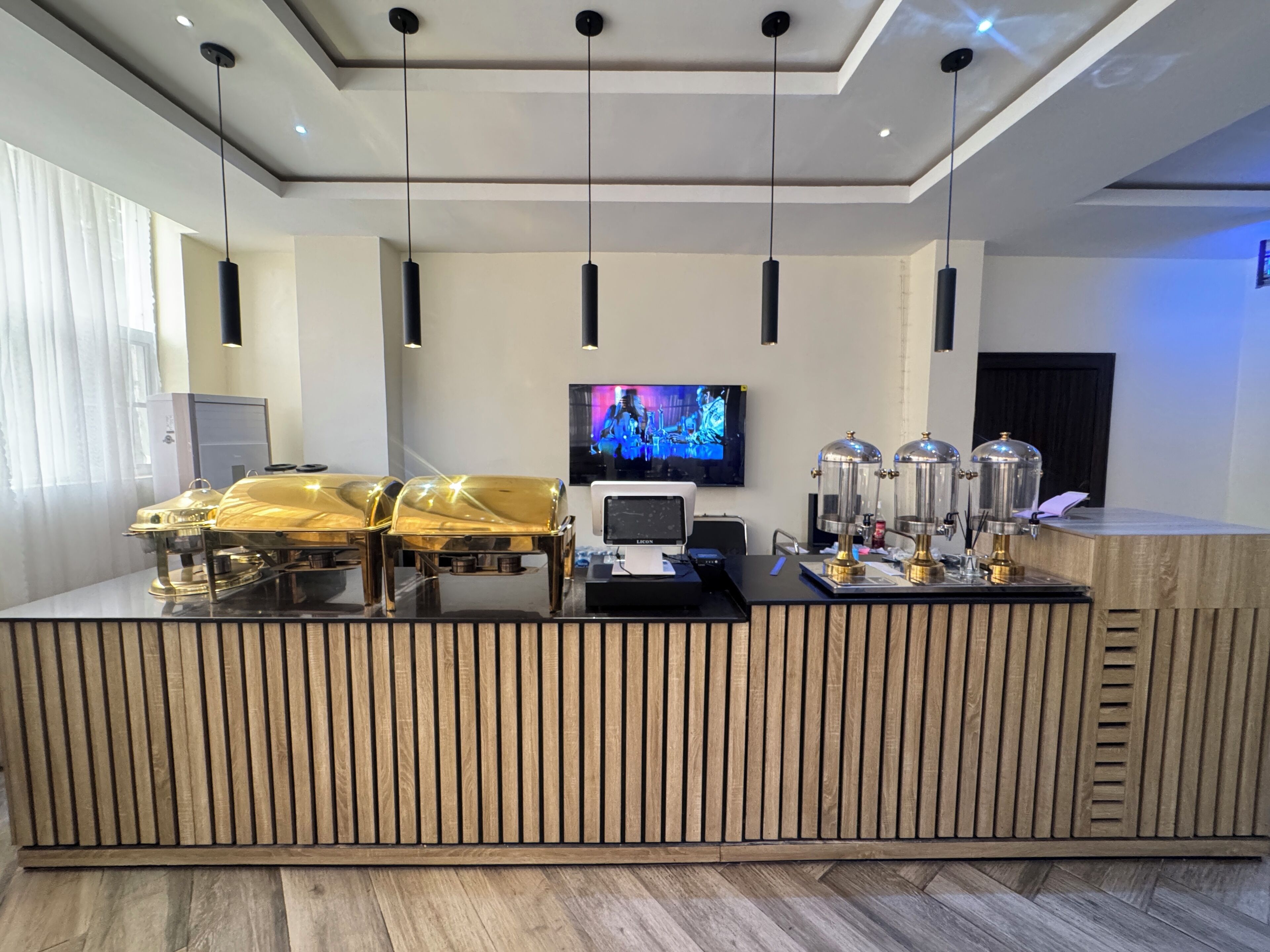 A reception desk with a modern design, featuring a wooden panel backlit with vertical lines, a flat-screen TV mounted on the wall, and a coffee machine.
