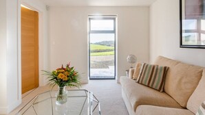 Un salón moderno con una mesa de centro de vidrio, un sofá color beige y vistas a un paisaje verde a través de un ventanal grande.