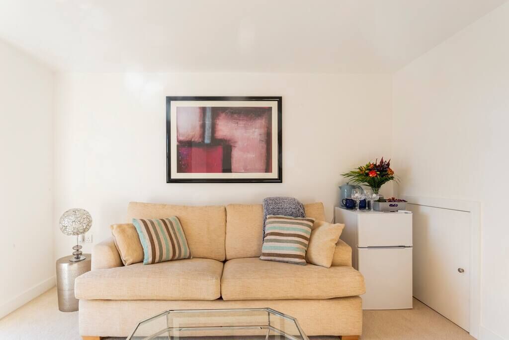 A living room with a beige sofa, a glass coffee table, a framed abstract painting, a small side table with a lamp, and a white cabinet with decorative items.