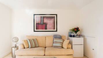 A living room with a beige sofa, a glass coffee table, a framed abstract painting, a small side table with a lamp, and a white cabinet with decorative items.