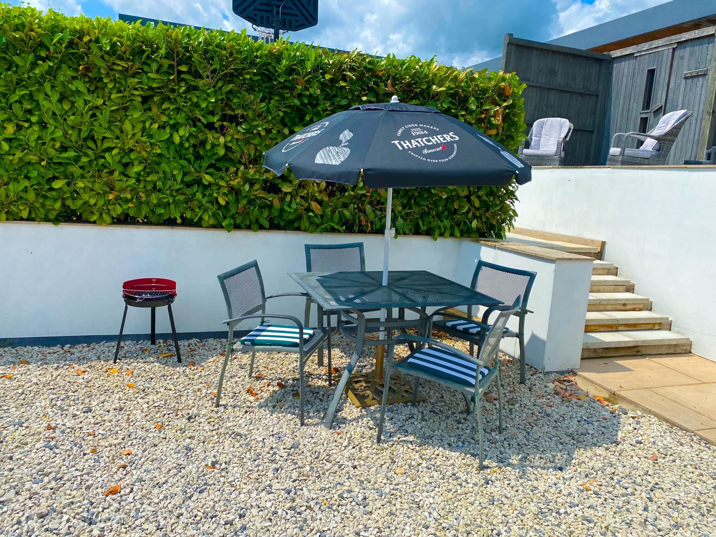 An outdoor patio with a table and chairs, a grill, and a hedge in the background.