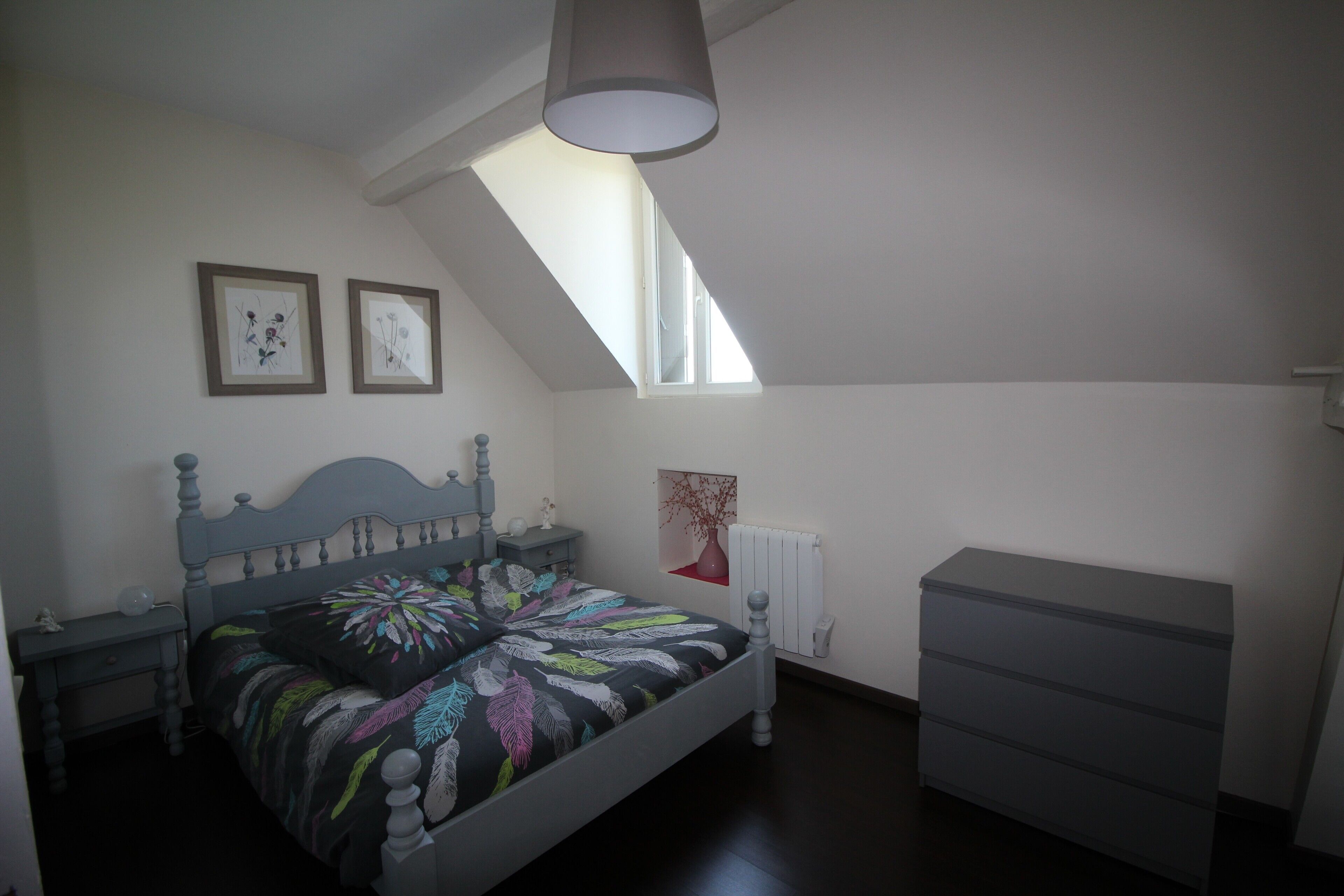 A bedroom with a sloped ceiling, a bed with a floral bedspread, a dresser, and two framed pictures on the wall.