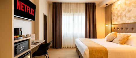 A hotel room with a bed, a desk with a computer, a TV displaying 'NETFLIX', and a window with curtains.
