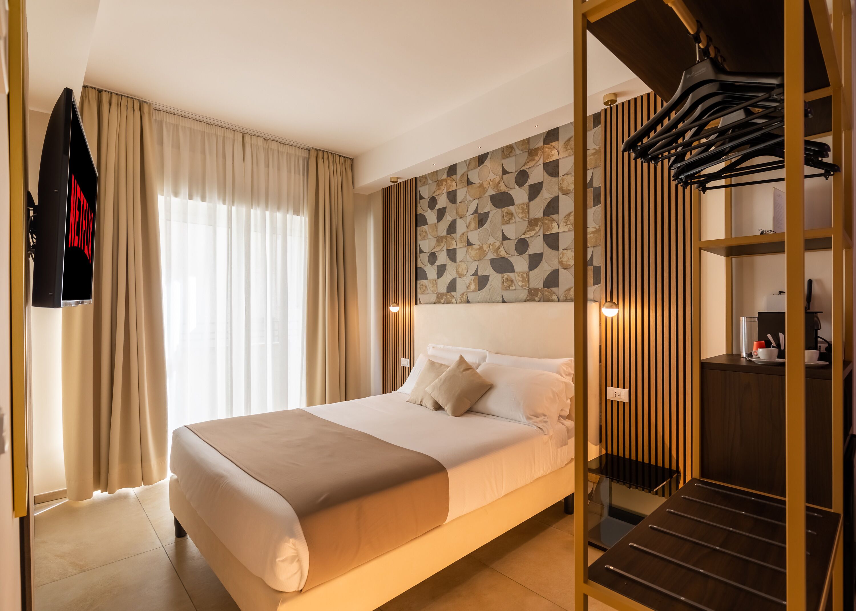 A modern hotel room with a large bed, a wardrobe with a mirror, a desk with a chair, and a view of the outside through large windows.