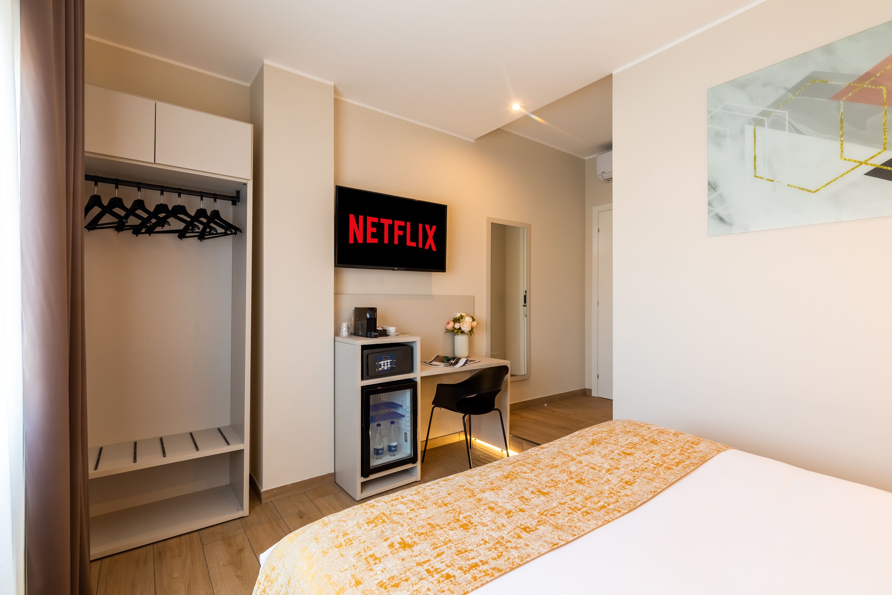 A modern hotel room with a bed, a desk with a computer, a mini-fridge, and a coat rack.