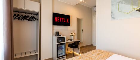 A modern hotel room with a bed, a desk with a computer, a mini-fridge, and a coat rack.