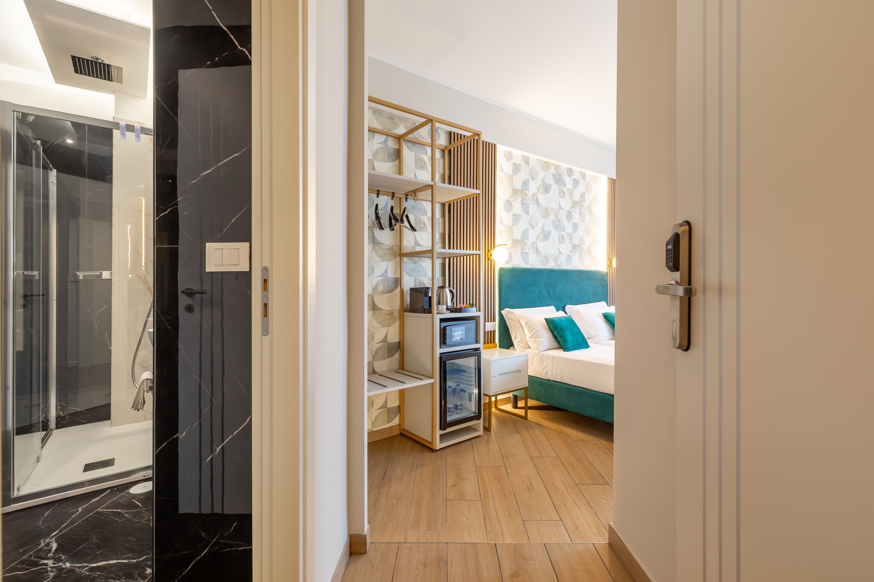 Una habitación de hotel moderna con ducha de vidrio, armario de madera, cama con ropa de cama color turquesa y pared con estampado geométrico.
