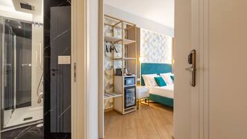 A modern hotel room with a glass shower, a wooden wardrobe, a bed with teal bedding, and a geometric-patterned wall.
