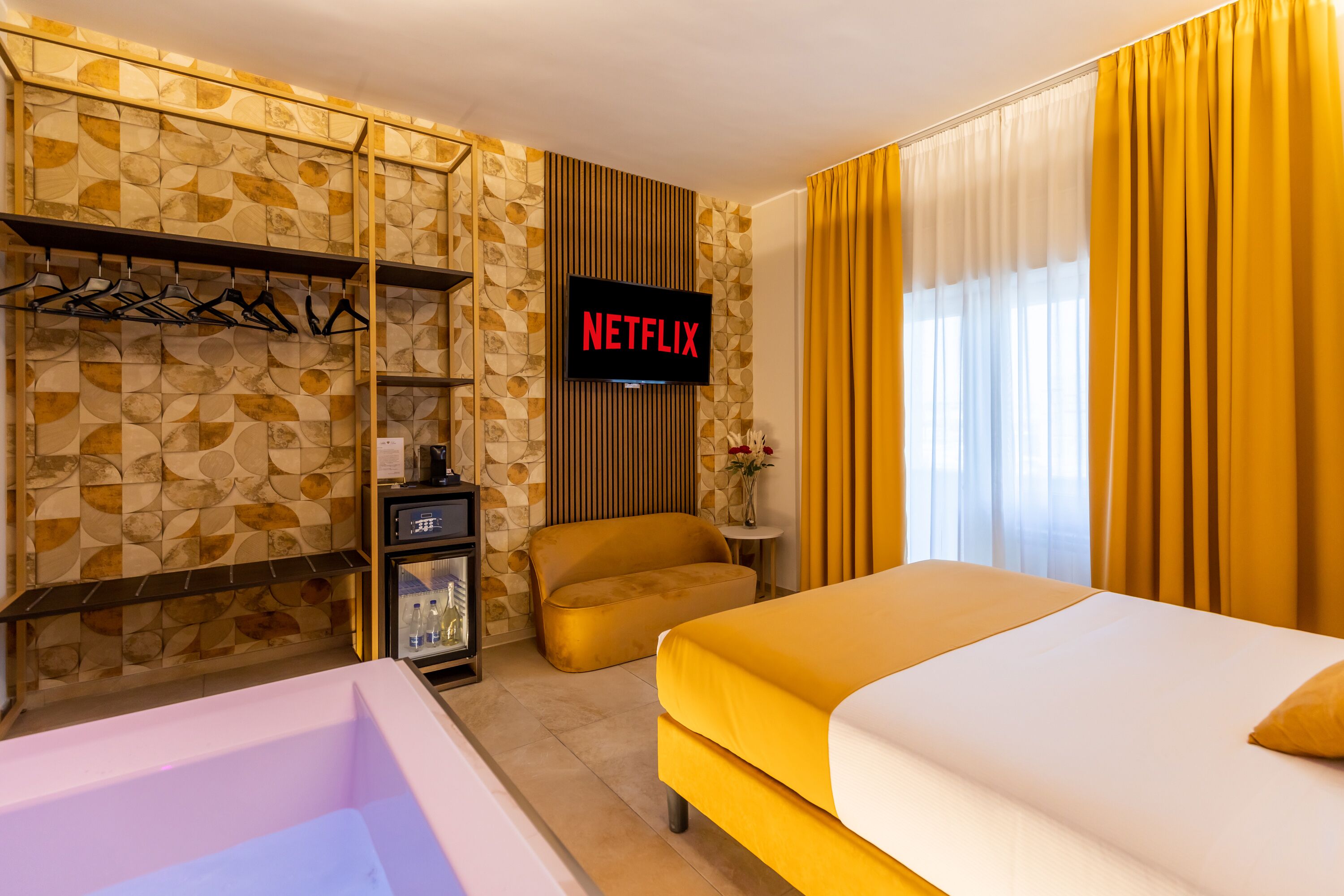A hotel room with a bed, a sofa, a TV displaying the Netflix logo, a minibar, and a wall with a geometric pattern.