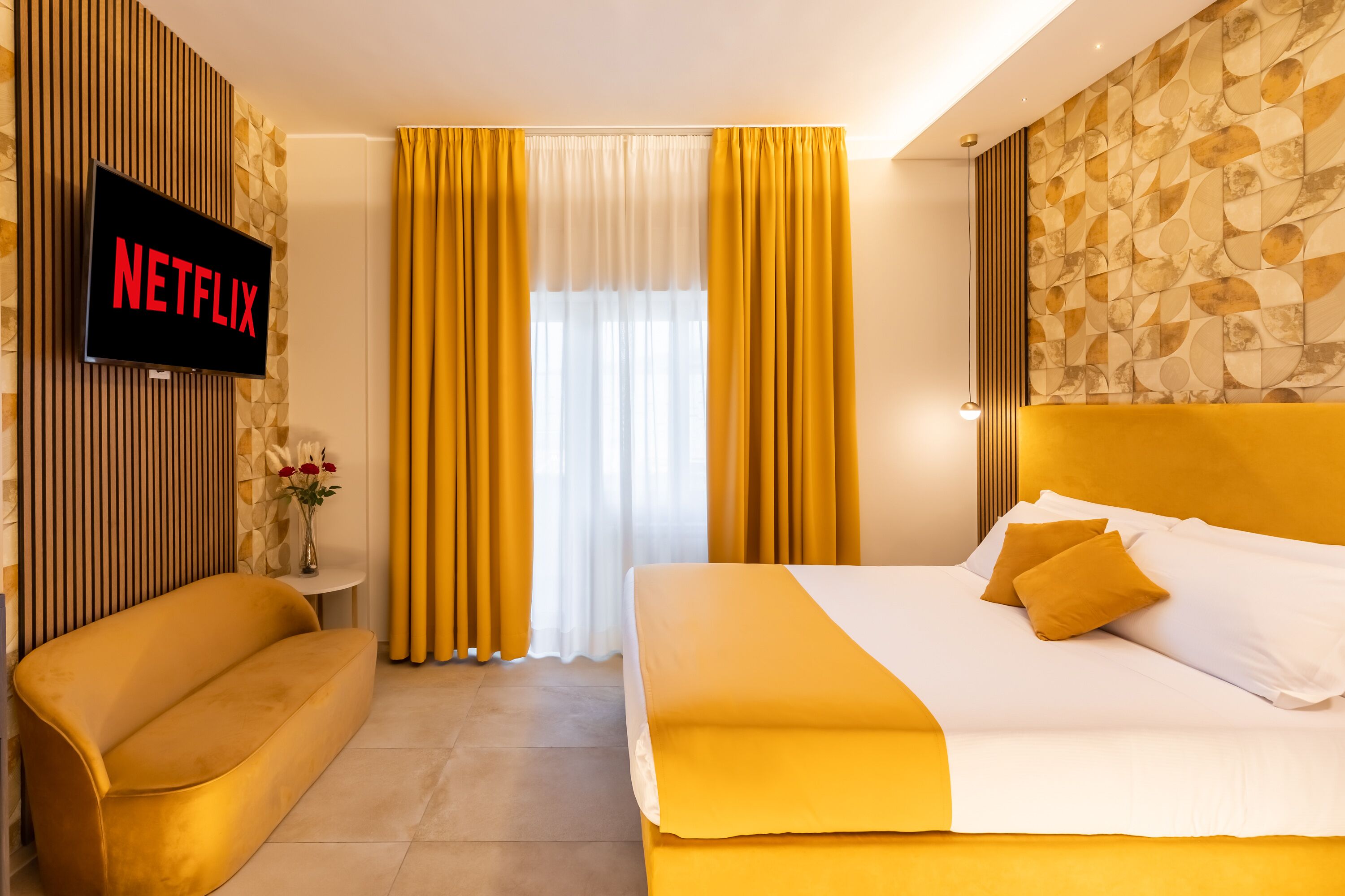 A hotel room with a bed, a sofa, a TV displaying 'NETFLIX', and yellow curtains.