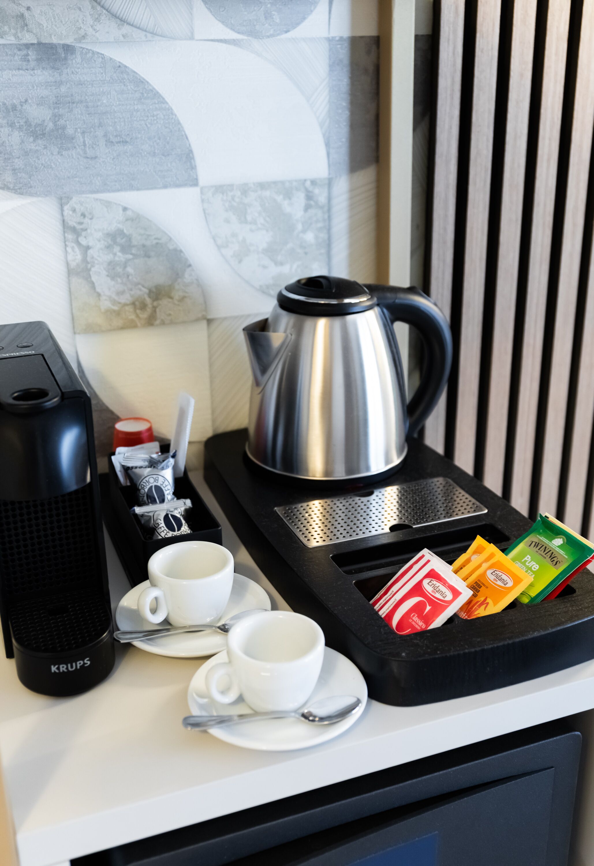 A coffee station with a Krups coffee maker, a silver electric kettle, and two white cups with saucers.