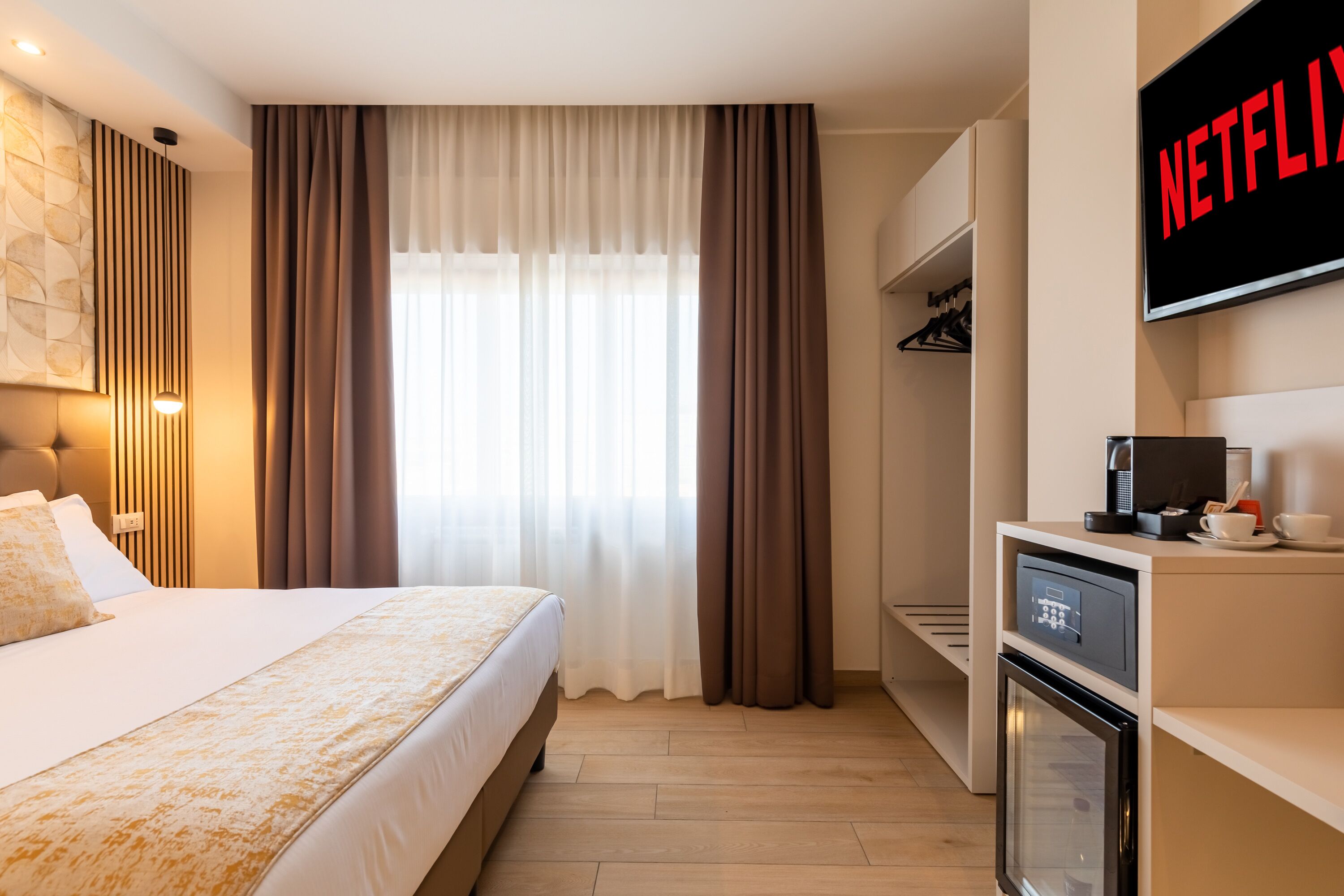 A modern hotel room with a bed, a TV mounted on the wall, a minibar, and a built-in oven.