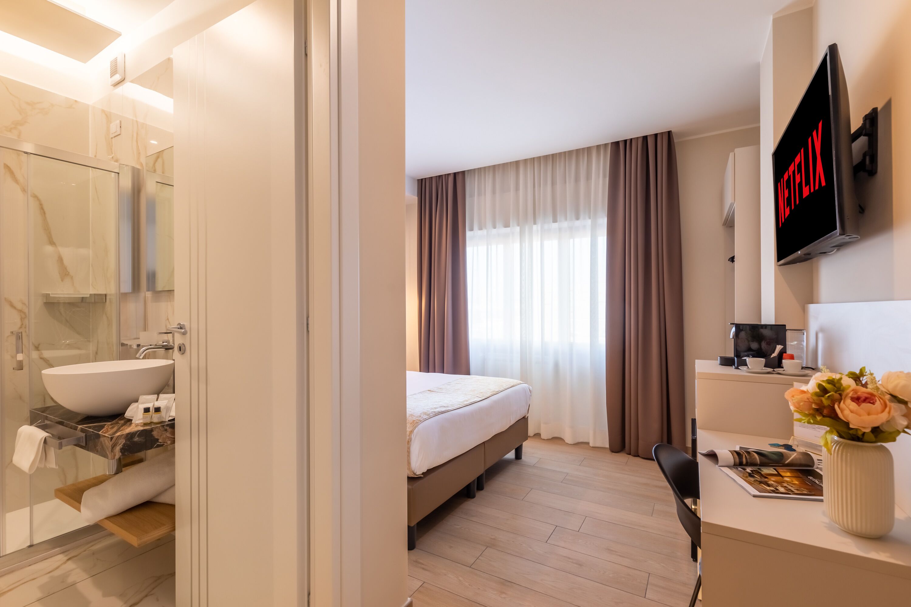 Een moderne hotelkamer met een groot bed, een bureau met een bloevas, een bevestigde televisie met het Netflix-logo en een badkamer met een grote spiegel, wastafel en een marmeren achterwand.