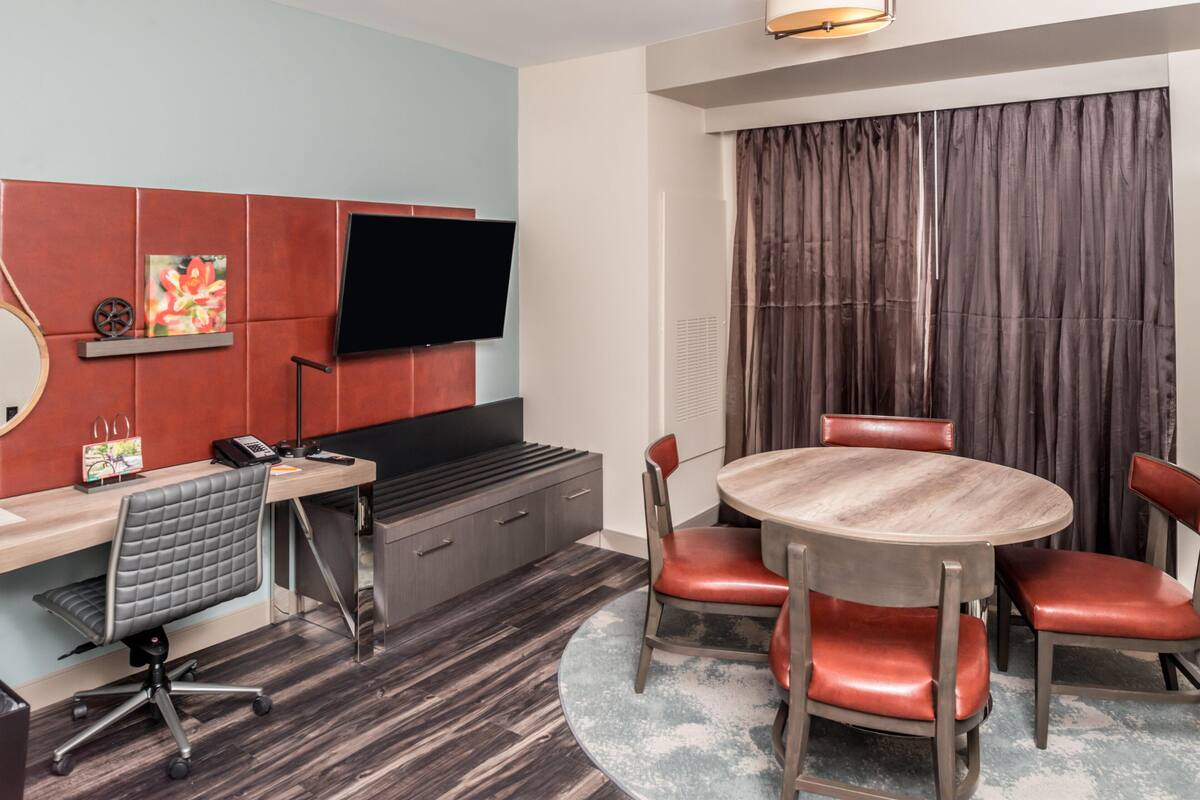 A hotel room with a flat-screen TV, a desk with a chair, a round table with chairs, a large window with curtains, and a wooden floor.