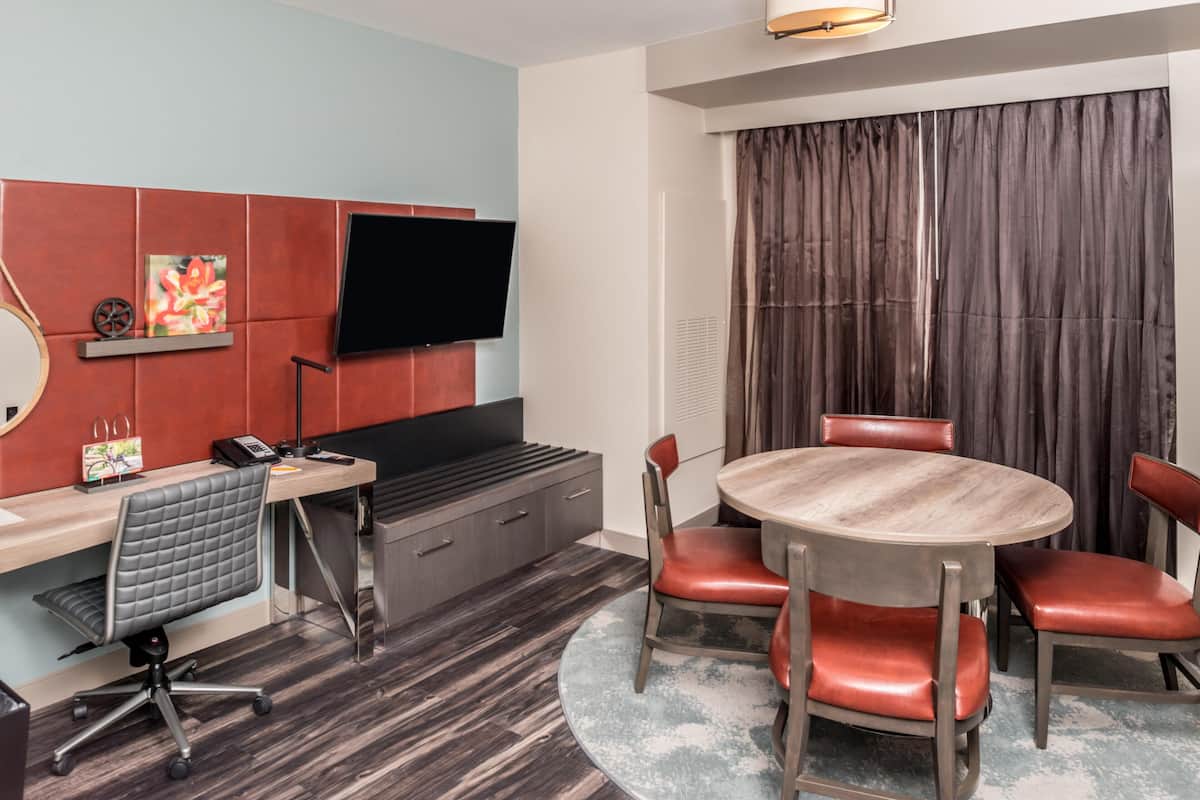 Hotellihuone, jossa on litteänäyttöinen televisio, työpöytä tuolilla, pyöreä pöytä tuoleineen, suuri ikkuna verhoilla ja puulattia.