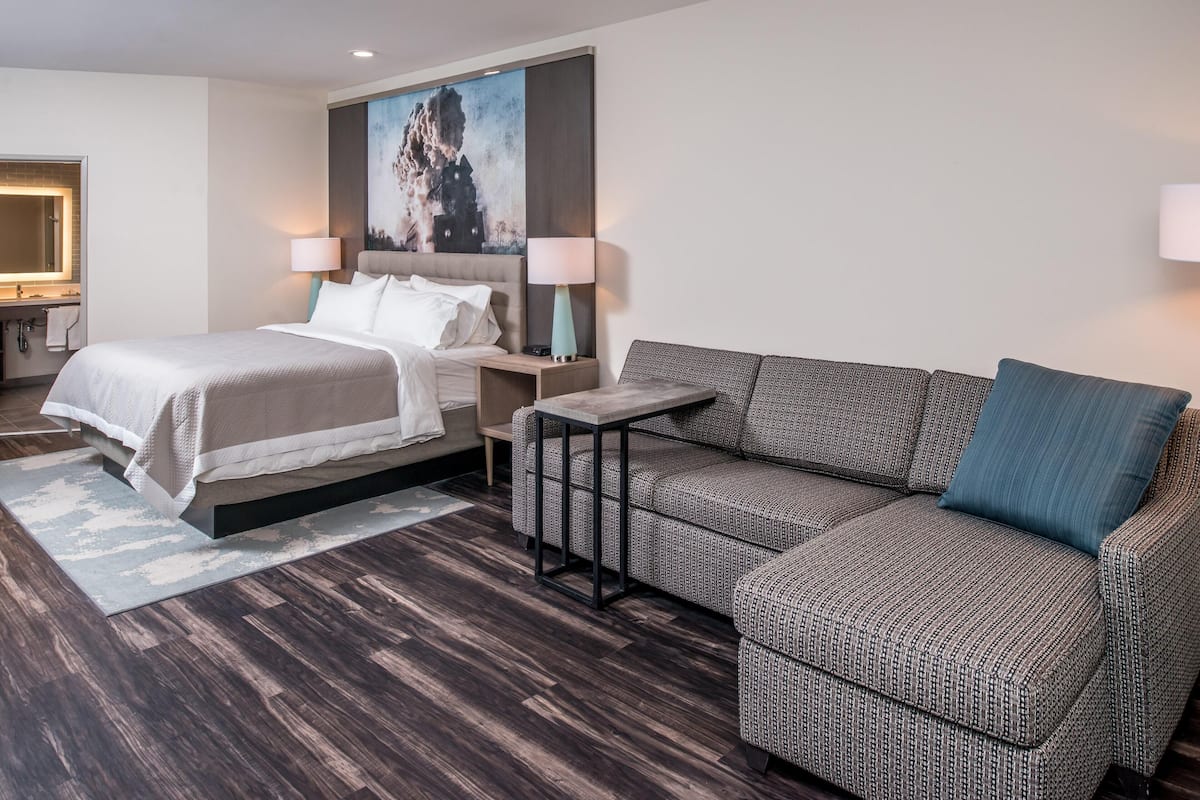 A modern hotel room with a bed, a sofa, a nightstand, a lamp, and a painting on the wall.