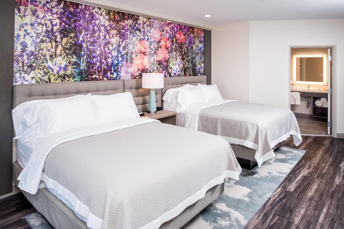 A hotel room with two beds, a large floral artwork on the wall, a bedside table with a lamp, and a bathroom visible through an open door.