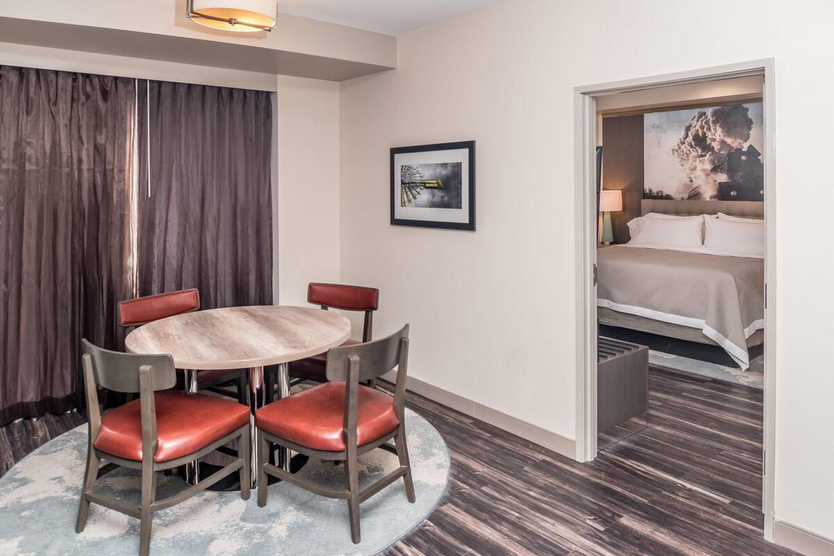 A hotel room with a round table, four chairs, a bed, and a painting on the wall.