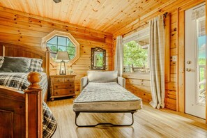 A wooden cabin room with a bed, a small table with a lamp, a mirror, and a window with curtains.