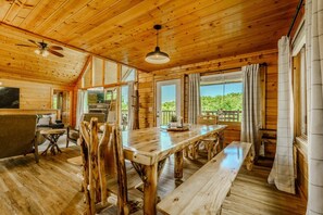 A wooden cabin interior with a dining table, chairs, a ceiling fan, and large windows.