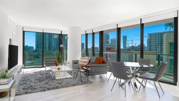 A modern living room with a gray sofa, a coffee table, and a dining table with chairs. Large windows offer a view of the city skyline and construction cranes. - HYDE MIDTOWN RESIDENCES SPECTACULAR CORNER 2BR (Miami)