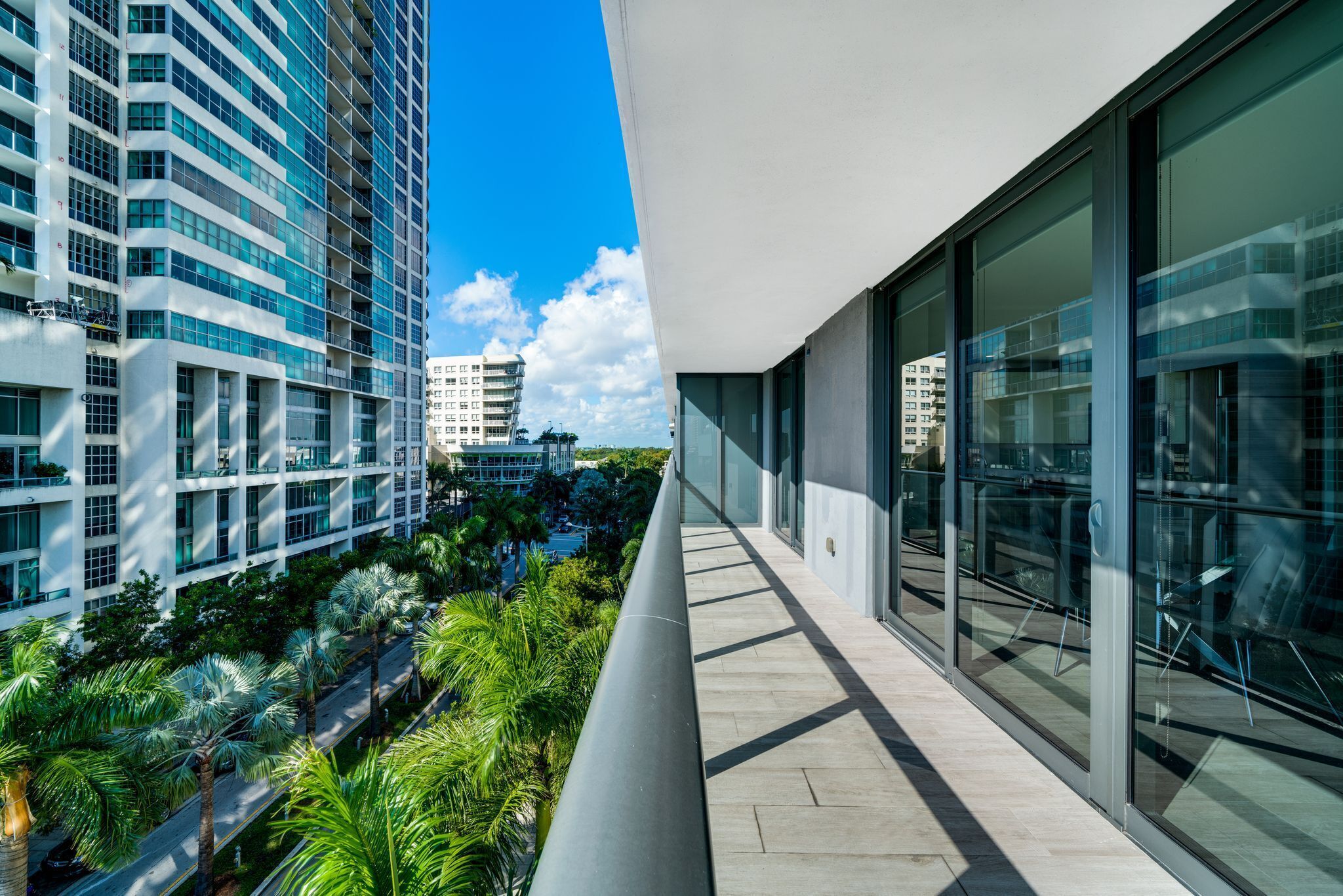 A modern building with a glass facade, a balcony with a view of palm trees and other buildings, and a clear blue sky.