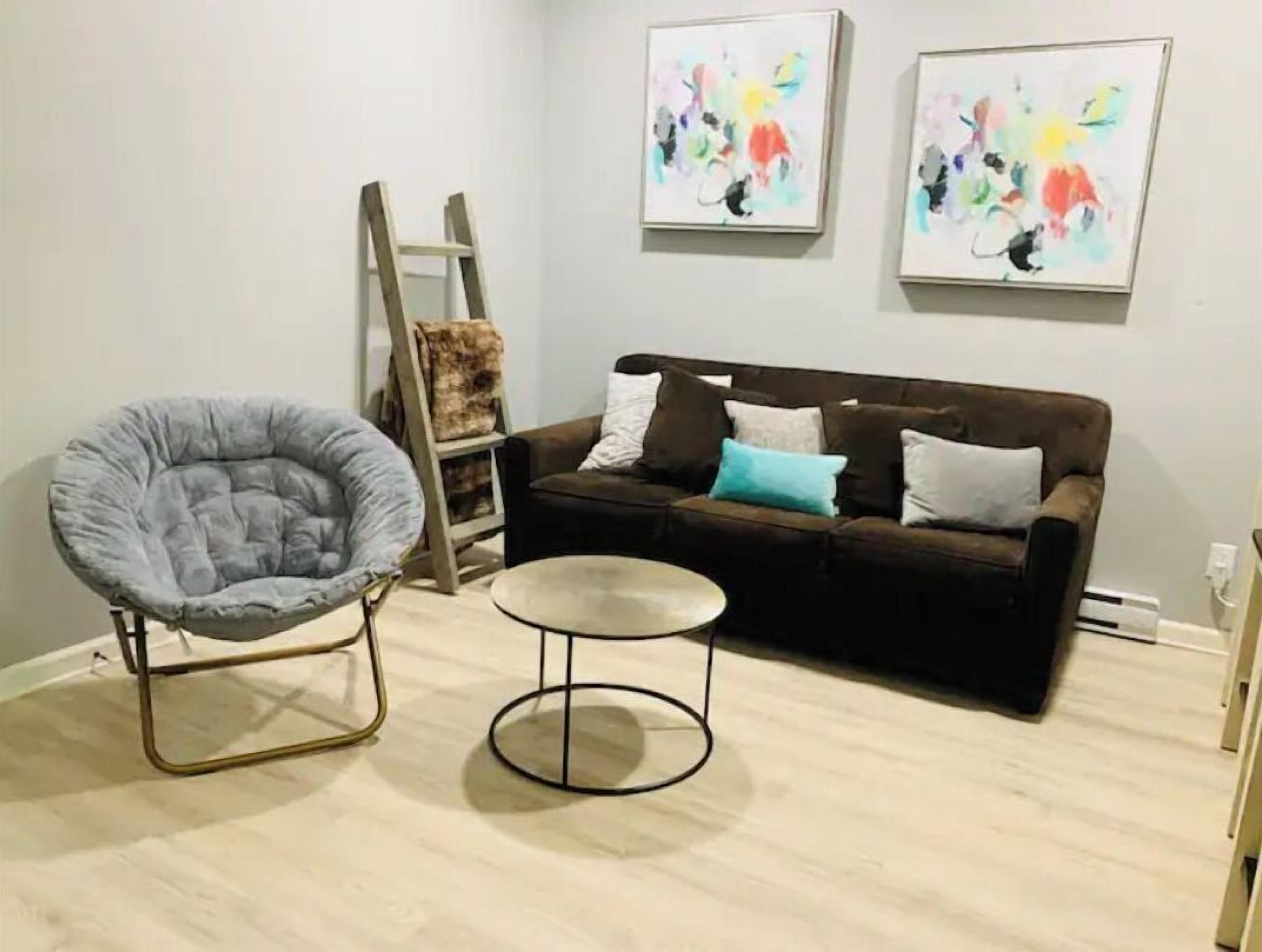 A living room with a grey chair, a brown sofa with cushions, a small round table, and two abstract paintings on the wall.