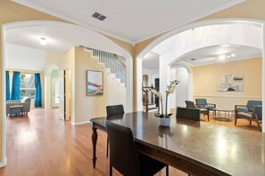 A spacious living room with a large wooden dining table, comfortable seating, a staircase, and a view of the outdoors.