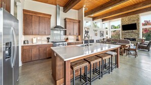A spacious kitchen with wooden cabinets, a large island with bar stools, a stone fireplace, and a TV mounted on the wall.