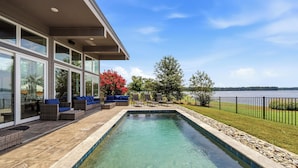 A swimming pool with a clear view of the lake, a covered patio with seating, and a modern house with large windows.
