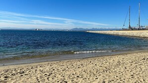 A sandy beach with a calm sea, a stone pier with sailboats, and distant mountains under a clear blue sky. - V021 - Neptuno (Roses)