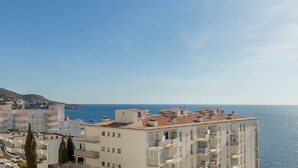 A coastal hotel with multiple balconies overlooking the sea, a clear sky, and a hilly landscape in the background. - V021 - Neptuno (Roses)