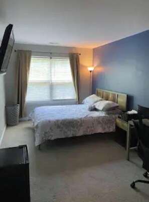 Cozy retreat with a single bed, desk, and chair, featuring a window with curtains, flat-screen TV, and lamp.