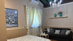 A bedroom with a bed, a sofa, a small table, a chandelier, a wall-mounted air conditioner, and a poster.